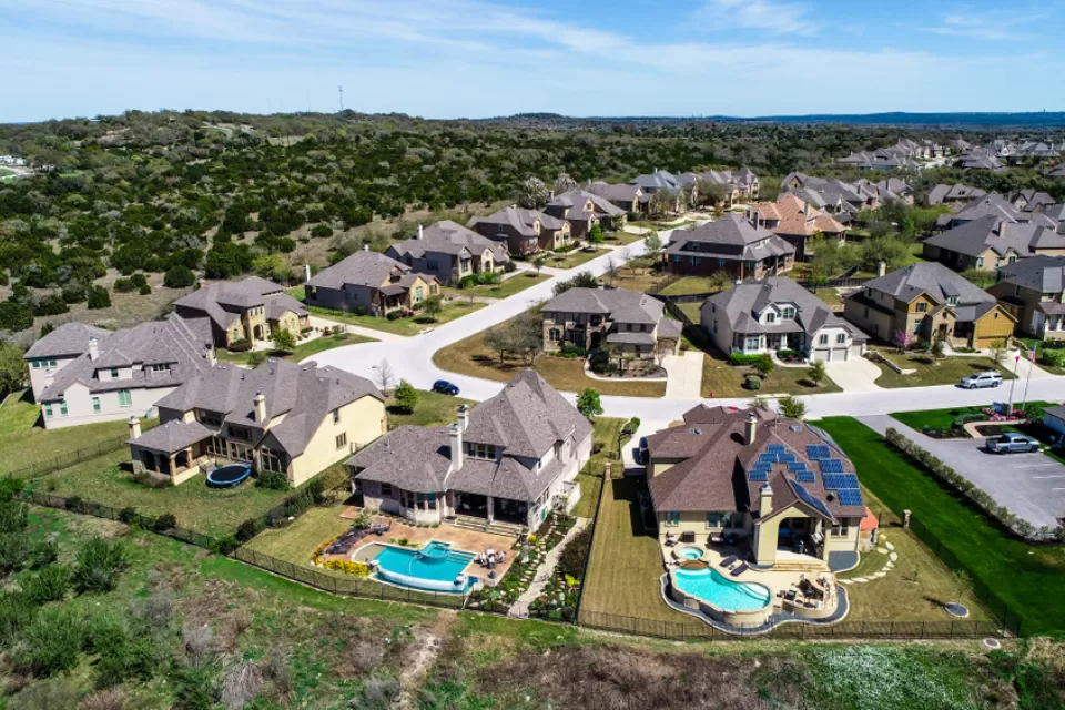 A subdivision of large suburban homes, some with bright blue pools, sits atop a low hill in Hays County, Texas.
